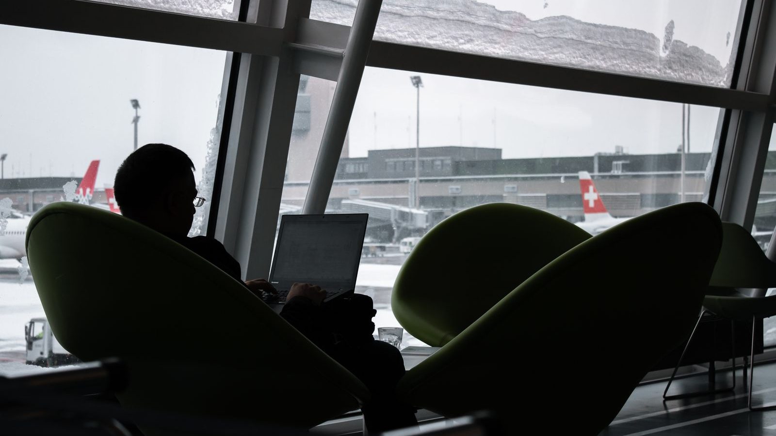 一名旅客坐在機場貴賓室窗邊的扶手椅上使用手提電腦，窗外雪地停機坪上停泊着客機。
